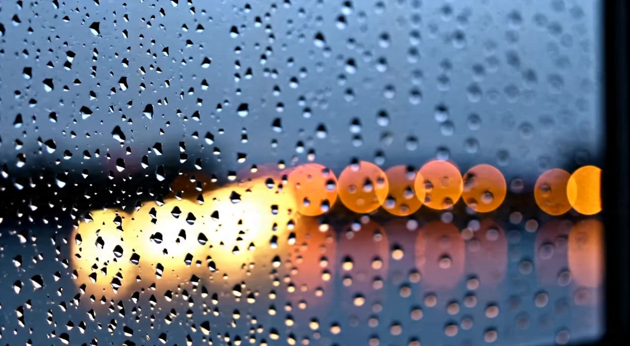 Rain droplets sliding down a window with glowing city bokeh beyond.