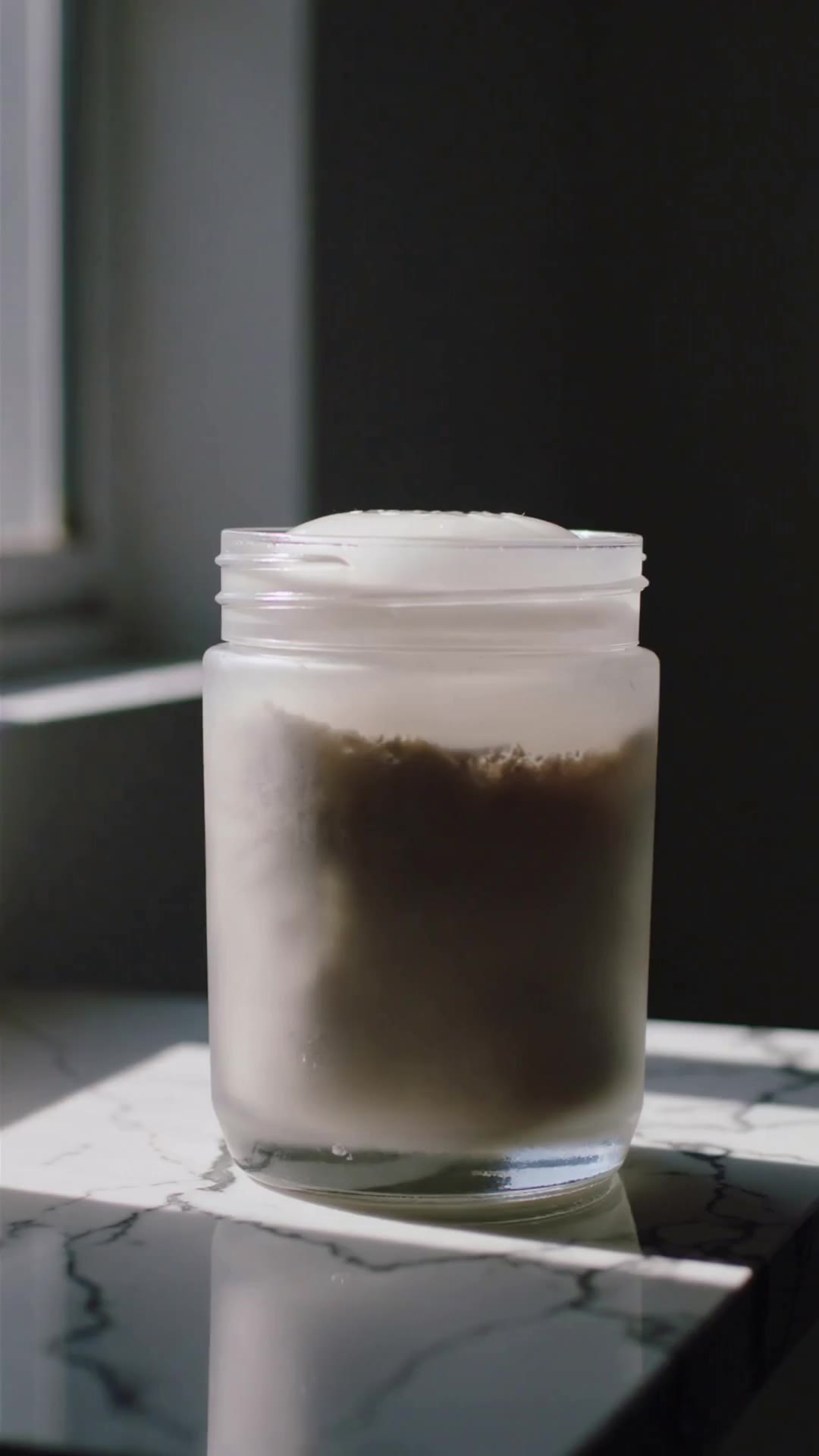 An unbranded frosted-glass skincare jar lifted from a marble counter in soft window light.